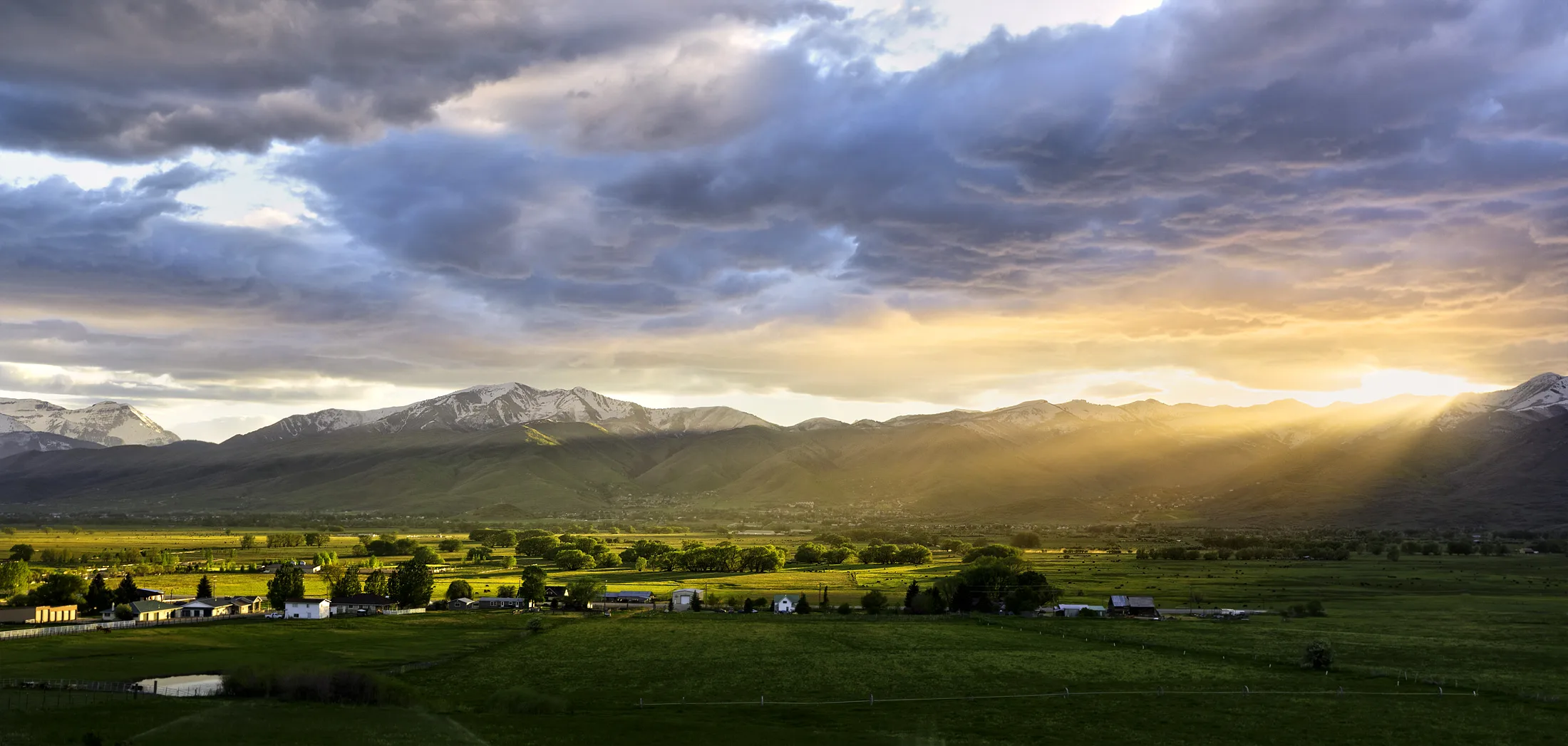 Evening sunlight breaks through storm clouds over the snow-capped mountains and green fields of Heber Valley